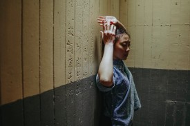 A woman stands with her hands above her head against a two-tone wall in a still from Within These Walls